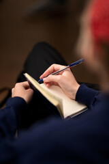 Person is writing with pen in notebook with his right hand indoors. Student in red hat is outlining...