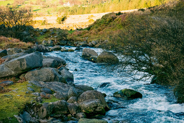 A tranquil river flows swiftly over large boulders surrounded by lush greenery. The tranquil setting features rolling hills under a clear sky, inviting relaxation and exploration.