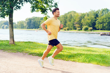 Man jogging along a serene riverside path on a sunny day surrounded by lush greenery
