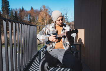 Young musician sits on a sunny balcony, immersed in playing the ukulele. Surrounded by autumn...