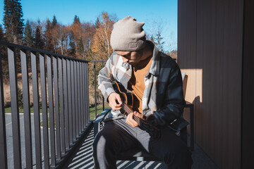 Young musician sits on a sunny balcony, immersed in playing the ukulele. Surrounded by autumn...