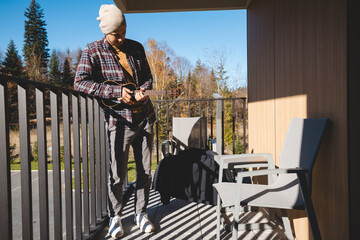 young musician stands on a sunlit balcony, playing the ukulele while gazing into the distance....