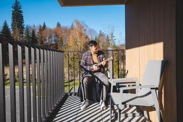 Young musician sits on a sunny balcony, immersed in playing the ukulele. Surrounded by autumn...