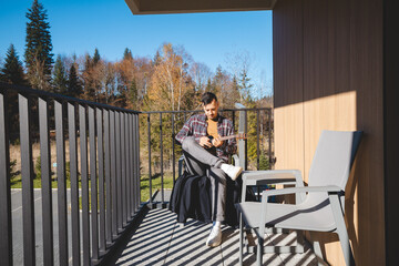 Young musician sits on a sunny balcony, immersed in playing the ukulele. Surrounded by autumn...