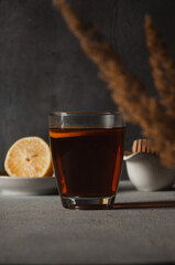 Glass with black tea and slice of lemon on the table, honey spoon and lemon in the background. Vertical photo. Breakfast and morning hot beverage concept. Dark mood