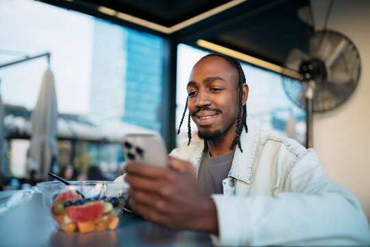 Smiling black man using smartphone and eating fruit salad at outdoor cafe