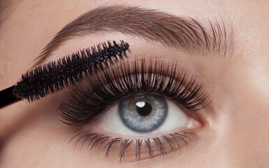 A close-up of a person's eye, emphasizing the eyelashes and eyelid. A mascara brush is applied to the eyelashes, making them appear longer and fuller. 