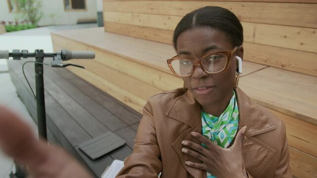 UGC handheld shot of young African American business lady in glasses and earphones talking emotionally to camera, gesturing while talking about new project outdoors, scooter standing behind her