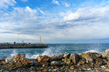 Waves Crashing on Rocks, Cloudy Sky, Rocky Coastline with Jetty and Seagulls or gulls. Yellow navigation light post or Lighthouse on the concrete pier. Waterfront View with Seabirds