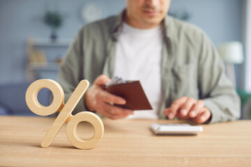 Soft focus closeup of wooden percent sign on desk, with man holding his wallet and calculating...