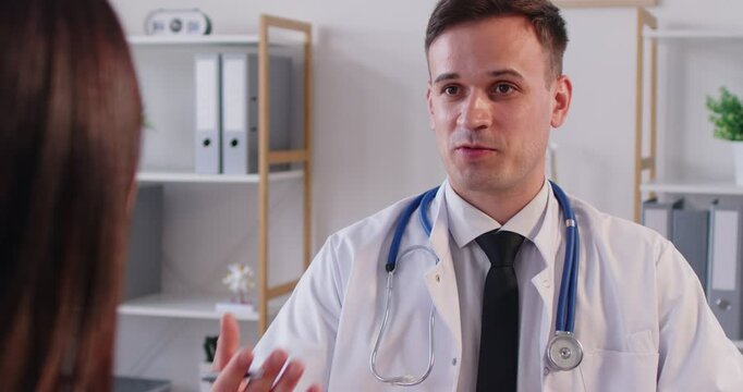 Young male doctor in white medical uniform talking to discuss, share results or symptoms with female patient, GP or physician consulting woman client to give recommendation at meeting in hospital