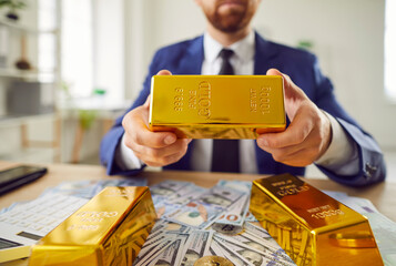 Rich man wearing business suit, holding a solid gold bar, surrounded by cash on table, symbolizing...