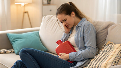 Fototapeta premium Young Woman Holding a Red Hot Water Bottle to Her Chest While Experiencing Emotional Menstrual Pain at Home