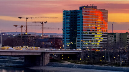 A modern cityscape captures the festive spirit of Christmas with a building's lights reflecting a giant holiday tree against a backdrop of construction cranes at sunset