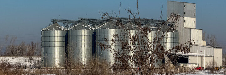 A row of metal grain silos stands amidst a snowy landscape, representing agricultural storage solutions and winter harvest challenges