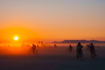 A warm orange sunset illuminates a desert festival scene with cyclists, art installations, and...
