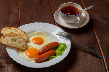 Breakfast fried eggs with sausages, fresh croutons, and tea in a cup on a brown wooden table