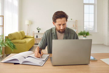 Fototapeta premium Portrait of confident bearded young business man in casual clothes working at the desk on his workplace with laptop at office or at home making notes in his notepad looking at computer monitor screen