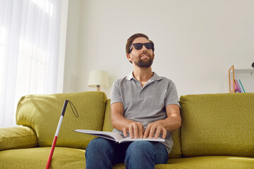 Portrait of a young bearded blind man wearing dark eyeglasses sitting on sofa in the living room at...