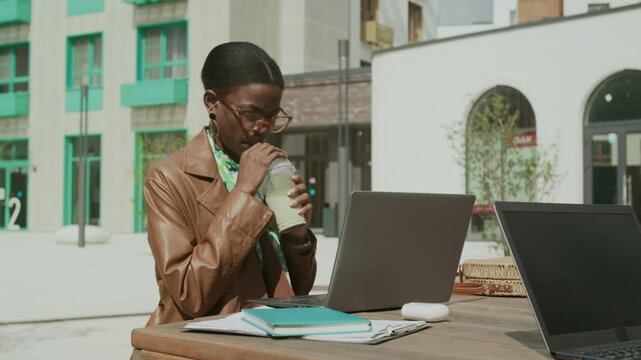 Medium shot of Black businesswoman in brown tench coat and glasses sitting at outdoor table in courtyard of modern house, drinking smoothie and working on laptop on sunny summer day
