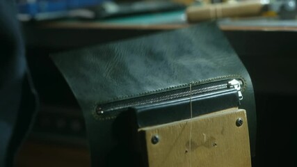 Craftsman is carefully sewing a zipper onto a piece of green leather, using a needle and thread. He is working on a wooden table in his workshop