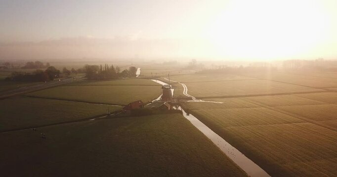Summer morning sun opposite. Drone moves from right to left. In the center of the frame is an old Dutch mill located on a green field near a canal with water. Province of Groningen. North Netherlands.