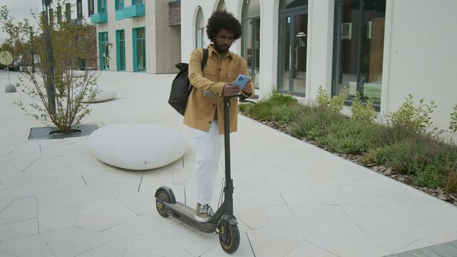 Full shot of young Biracial Businessman in yellow shirt standing on electric scooter in middle of residential courtyard, holding phone in his hands and reacting ambiguously to results of projects