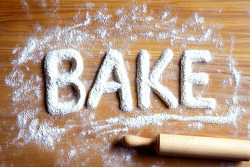 "BAKE" is written in flour on a wooden surface with a rolling pin. Concept of baking, kitchen, homemade, cooking.