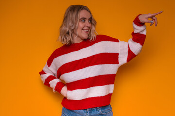 A smiling woman in a red and white sweater points to the side, standing against a vibrant orange...