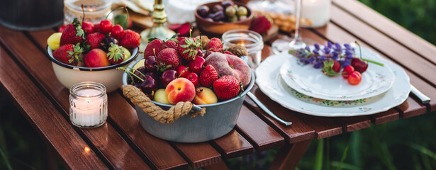 Banner. Elegant gorgeous wedding table decor or romantic dinner arrangement outdoors in blooming field. Purple lupine flowers, candles, fruits, wine, wooden furniture. Sunset, summer, golden hour