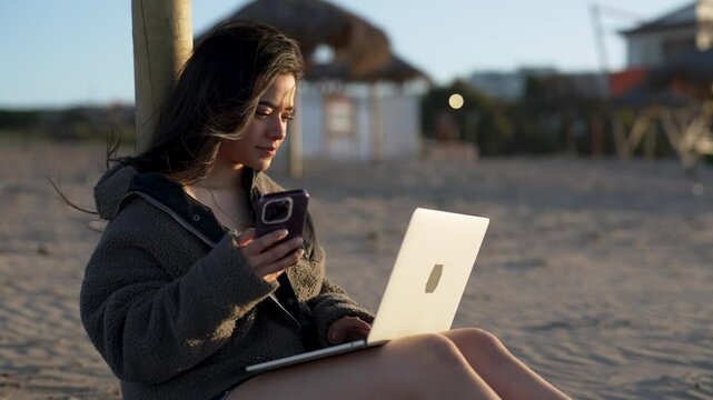 latin Digital nomad woman working using mobile phone and notebook on the beach at sunset