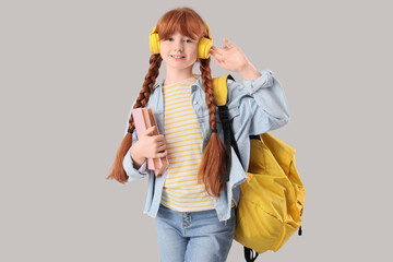 Teenage redhead girl in headphones with books and backpack on light background