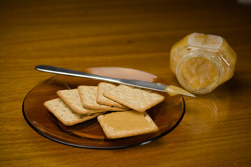 A plate of crackers topped with golden honey, accompanied by a jar of honey and a knife on a wooden table, offering a simple and sweet snack option.