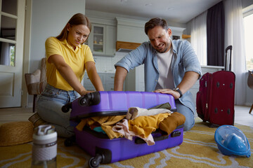 Young loving couple with frowning face trying to close overloaded travel bag