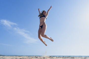 Mujer joven saltando de emoción porque esta feliz y divertida, con un sensacional día de vacaciones en verano en la playa, soleado con cielo azul.