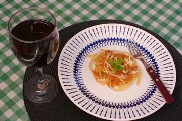  plate of linguine with tomato sauce, topped with grated cheese and basil, served alongside a glass of red wine on a green checkered tablecloth and black placemat.
