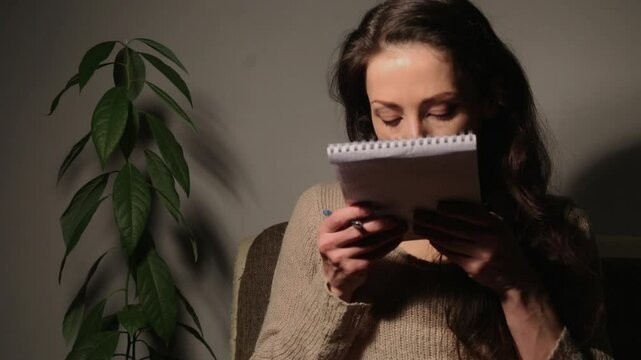 Happy laughing woman with cute smile jotting down notes with a pen in a note and kissing paper sits on sofa at home interioor near flower in studio light, low key, dark shadow. Emotion people closeup