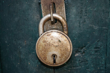 A close-up view of a circular, aged steel padlock secured to a latch on a weathered green wooden surface. The lock displays signs of wear and patina.