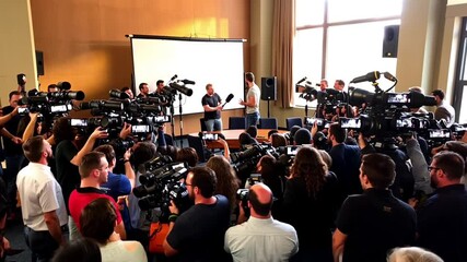 a man is holding a press conference in a room surrounded by many media crew covering it