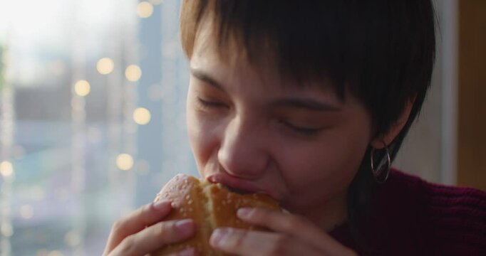 Close-up footage of a teenager girl eating burger while sitting at a cafe. This delightful video showcases a teenage girl savoring her burger, showcasing pure happiness and enjoyment.