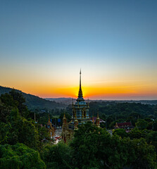Fototapeta premium A breathtaking aerial view of a golden Buddhist temple at Wat Mae Klang Waterfall in Chiang Mai Thailand is surrounded by lush greenery, with the sunrise casting warm hues across the horizon.