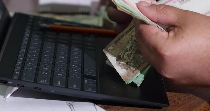 Person counts Hungarian forint cash notes next to laptop while seated at wooden desk in cozy workspace.
