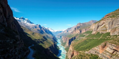 Fototapeta premium A winding mountain road follows a turquoise river through a dramatic canyon, nestled between towering cliffs and snow-capped peaks under a vibrant blue sky.