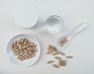 Capsule pills in a small white plate and in a white spoon, as well as a glass of water on a white background. Top of view. 