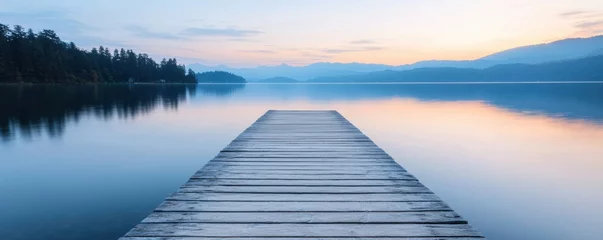 Fotobehang A serene wooden pier extends into a calm lake, reflecting soft hues of dawn while surrounded by tranquil mountains and lush trees. © Watcharaphon