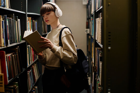 Young student in wireless headphones reading book near shelves