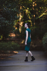 A woman walks confidently down a path in a lush green forest, wearing a dark green dress and black boots. Natural sunlight filters through the trees, creating a serene and vibrant atmosphere