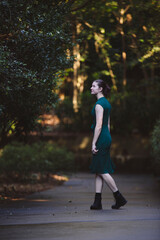 Obraz premium A woman walks confidently down a path in a lush green forest, wearing a dark green dress and black boots. Natural sunlight filters through the trees, creating a serene and vibrant atmosphere