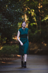 A woman walks confidently down a path in a lush green forest, wearing a dark green dress and black boots. Natural sunlight filters through the trees, creating a serene and vibrant atmosphere