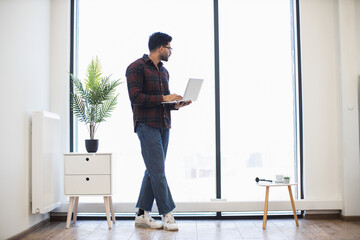 Young Indian male standing by large window with laptop in modern office. Casual attire with flannel shirt and jeans. Bright and minimalist workspace with indoor plant and natural light.